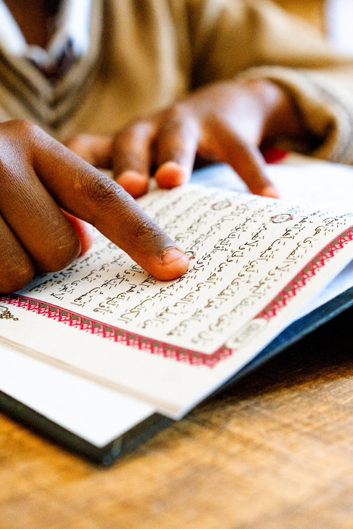 services-02 Close-up of a child's hand pointing while reading the Quran indoors.