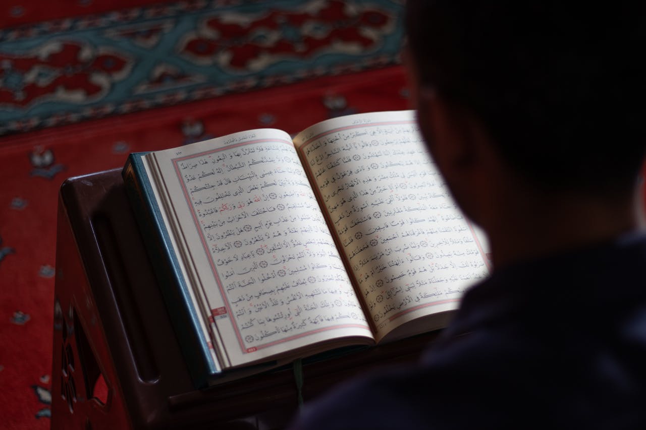 hero-img-01 A person reads the Quran inside a mosque with intricate carpet design, reflecting peace and spirituality.