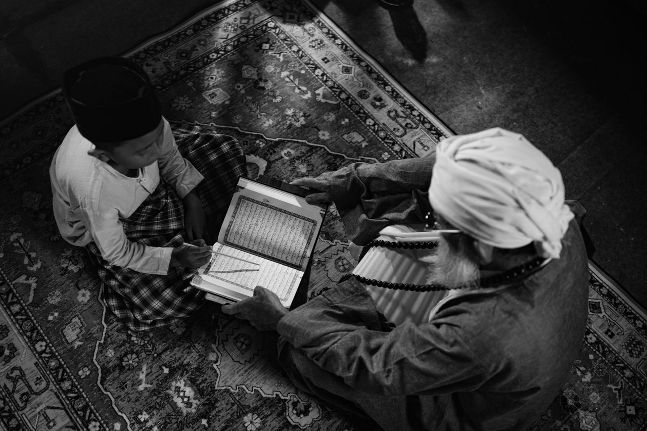 gallery-1 Black and white image of a boy learning from an elder, highlighting traditional Islamic education in Jakarta.
